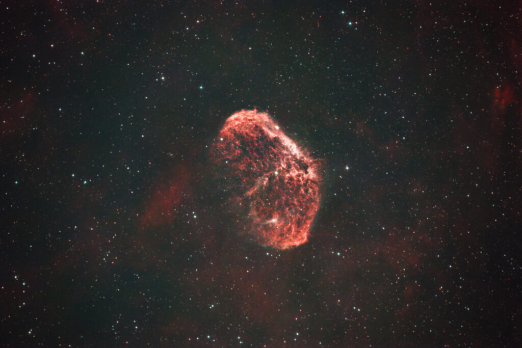 The Crescent Nebula glowing red against a dense field of stars and faint surrounding nebulosity.